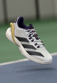Hvid atletisk sko med lilla accenter, der har tre sorte striber, en tekstureret overdel og en guld detalje på hælen. Designet til ydeevne.