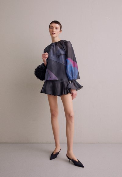 Blouse noire transparente avec des motifs géométriques colorés, jupe évasée noire ajustée et escarpins noirs à bouts pointus avec des accents en dentelle.