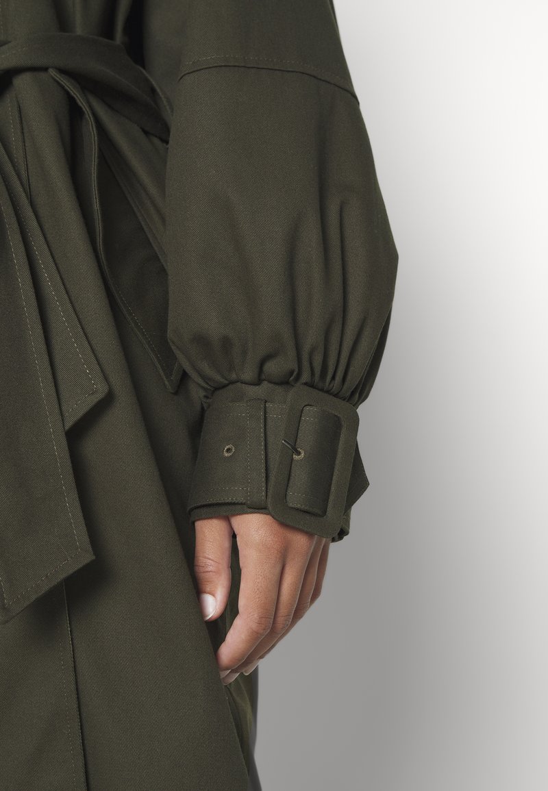 Olivgrüner Trenchcoat mit gepufften Ärmeln und einem Gurtverschluss, ausgestattet mit einer glatten Textur und sichtbaren Stichdetails.