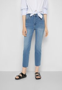 Jeans de mezclilla azul claro con pierna ajustada, recortados en el tobillo. Combinados con sandalias negras con dos correas ajustables.