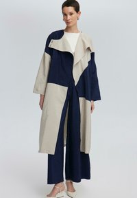 Lange jas in marineblauw en beige, met een losse fit, asymmetrisch ontwerp, brede mouwen en een gedrapeerde kraag. G dragen over bijpassende marineblauwe broek.