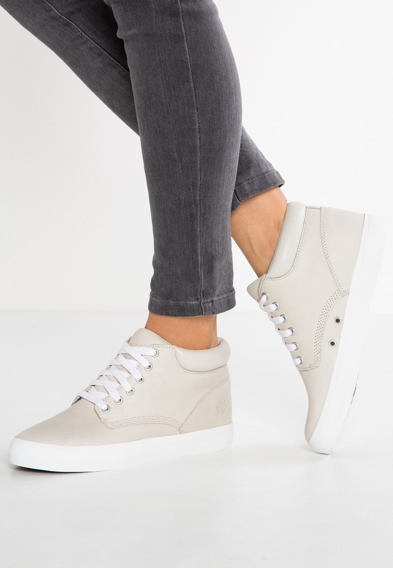 Personne portant des baskets montantes beige clair avec des lacets et semelles blanches, associées à un jean slim gris foncé sur fond blanc.