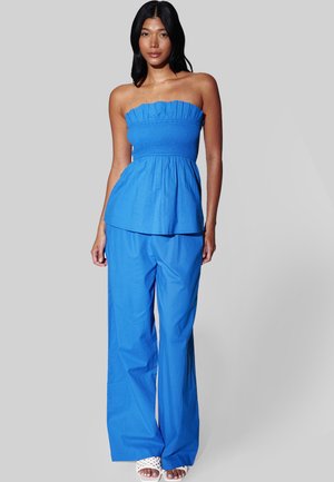 Vrouw draagt een blauw strapless top met plooien en bijpassende wijde broekspijpen, gecombineerd met witte open schoenen met geweven details, staand tegen een effen achtergrond.