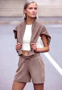 Brūns apgrieztais žakets, uzlikts uz pleciem, kopā ar baltu pieguļošu augšu un atbilstošām brūnām šortiem. Gluda tekstūra, ikdienišķs sportiskais dizains.