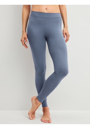 Vrouw met strakke blauwe legging staat op blote voeten met één been gekruist over het andere tegen een effen lichte achtergrond.