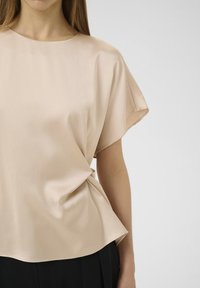 Top en satin beige avec des manches courtes et larges, et une coupe décontractée ; texture lisse et col arrondi ; porté avec un pantalon noir.