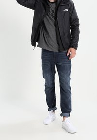 Men's outfit featuring a black, zippered jacket, gray t-shirt with logo, blue jeans, and white sneakers. Casual style, relaxed fit.