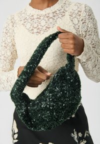 Vrouw in een crèmekleurige kanten top houdt een donkergroene paillettenhandtas vast met een rits bovenaan.