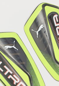 Φανταχτο κίτρινο νέον και μαύρες προστατευτικές επενδύσεις κνήμης Puma με μερικά κόκκινα γράμματα σε λευκό φόντο.