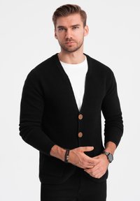 Černý pletený kardigan s výstřihem do V, dvěma dřevěnými knoflíky a žebrovanými manžetami. Nositelný přes bílý tričko s texturovaným povrchem.