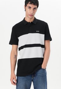 Svart og gråstripet polo skjorte med krage, med en liten hvit logo på brystet og korte ermer. Brukt med blå jeans.