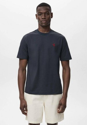 Dark navy t-shirt made of cotton with short sleeves and a crew neck; features a small red logo on the left chest. Worn with light beige shorts.