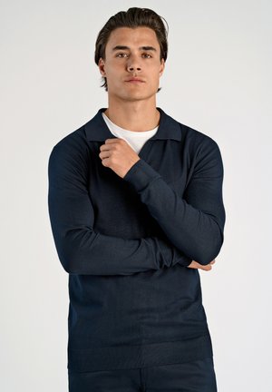 Suéter de polo de manga larga en azul marino, fabricado en tejido de punto fino, con cuello plano. Puños y dobladillo acanalados; usado sobre una camiseta blanca.