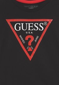 Svart T-shirt med en röd triangellogo som har texten "GUESS U.S.A." och ett frågetecken; inkluderar vit text och siffrorna 1201, 1203.