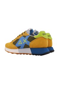 Zapatillas de ante amarillas y azules con acentos verdes, logo geométrico y suela blanca y azul en contraste. El diseño con cordones presenta múltiples texturas.