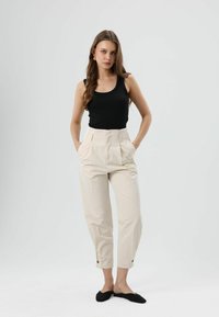 Schwarzes Tanktop, cremefarbene, plissierte, hochtaillierte Hose mit Knopfdetails, kombiniert mit schwarzen Slippern, steht vor neutralem Hintergrund.