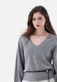 Grauer V-Ausschnitt-Pullover mit geripptem Muster, lockeren Ärmeln und einer taillierten Passform, akzentuiert durch eine braune Schnur mit metallischen Details.