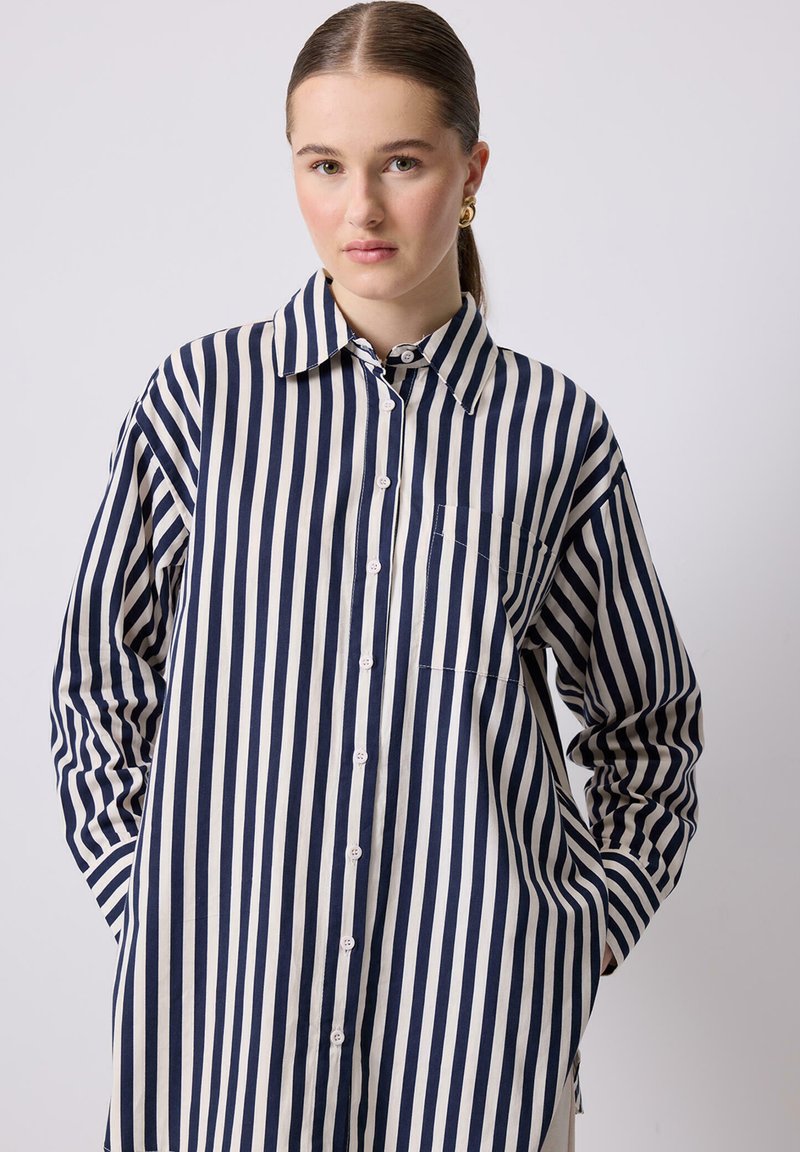 Camicia button-up con maniche lunghe, realizzata in tessuto leggero, caratterizzata da strisce verticali blu navy e crema, con orlo curvo e taschino sul petto.