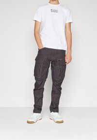 Bílé tričko s krátkým rukávem a černým logem, doplněné tmavě šedými nákladními kalhotami s mnoha kapsami a světle šedým prošíváním. Bílé tenisky.