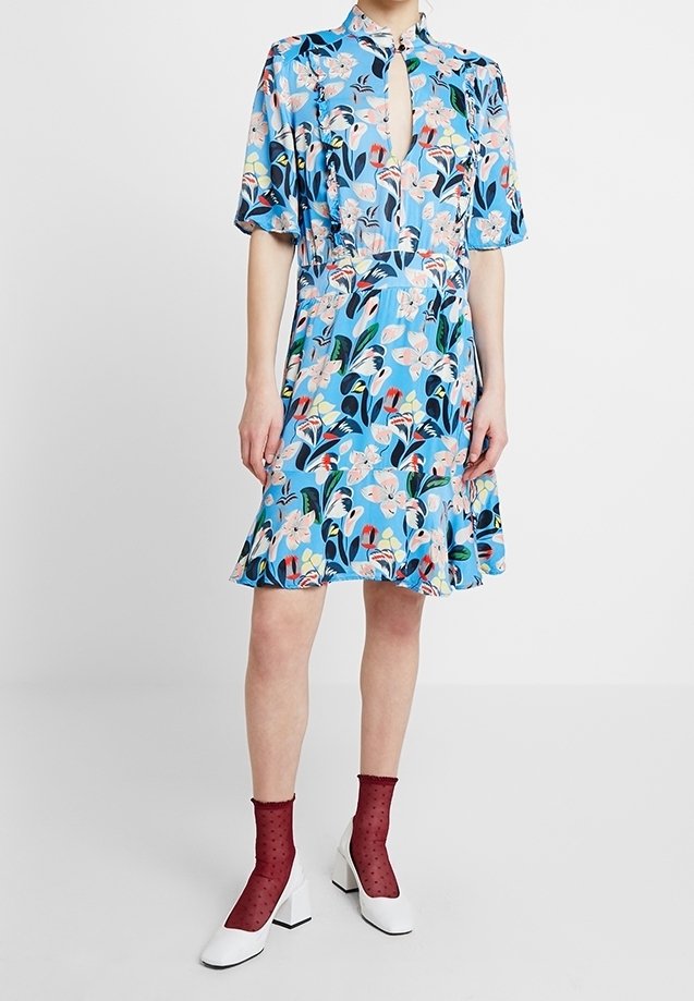 Femme portant une robe bleue à fleurs avec des manches bouffantes courtes, un décolleté en trou de serrure, et des chaussures blanches associées à des chaussettes rouges transparentes aux chevilles.