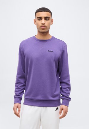 Giovane uomo con capelli corti che indossa una felpa viola girocollo con la scritta "BORG" e pantaloni bianchi con un piccolo logo nero.