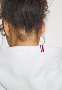 Felpa bianca con una finitura testurizzata, caratterizzata da un piccolo accento rigato rosso, bianco e blu nella parte posteriore del collo. L'immagine si concentra sul retro.