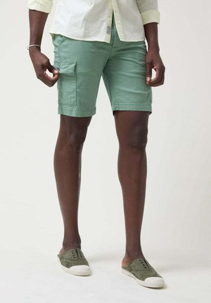 Pantalones cortos de carga verde claro con un frente plano y dos bolsillos laterales. Combinados con zapatos casuales verdes oliva. Tejido de algodón, corte relajado.