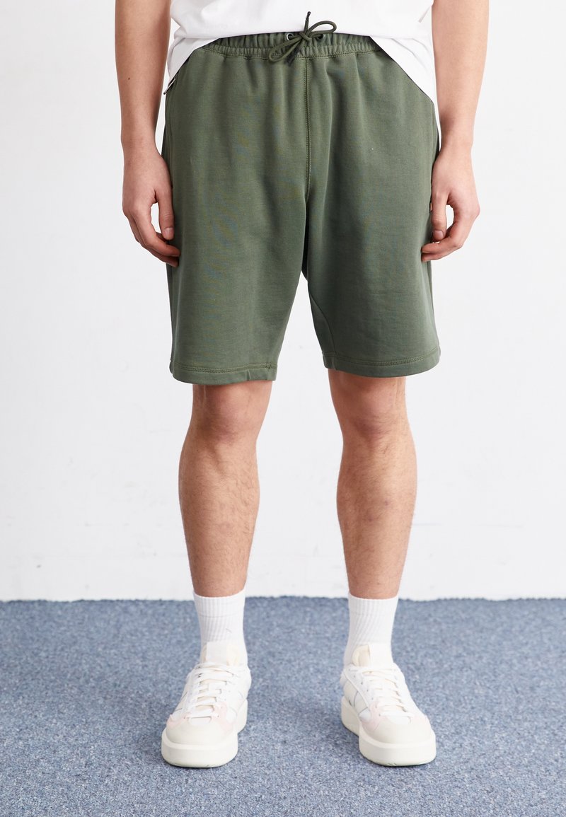 Homme debout portant un short vert olive à cordon, des chaussettes blanches et des baskets blanches sur un sol moquetté bleu.