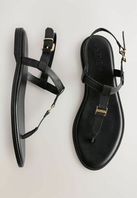 Sandalias planas negras con un diseño minimalista, que cuentan con correas de cuero, un separador de dedos y un acento de hebilla dorada en la correa del tobillo.