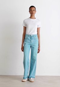 Hvit bomulls t-skjorte med lite logo, lyseblå høytlivs vide jeans, hvite og brune sneakers, stående mot en ensfarget bakgrunn.