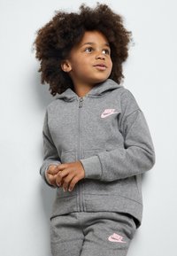 Sudadera gris con cremallera hecha de material de forro polar con un logo de Nike en rosa. Cuenta con una capucha y bolsillos laterales. Combinada con pantalones grises a juego.