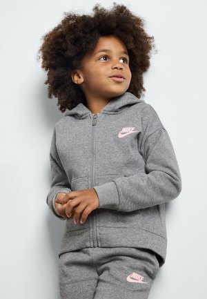 Haut à capuche zippé gris en matériau polaire avec un logo Nike rose. Comprend une capuche et des poches latérales. Associé à un pantalon gris assorti.