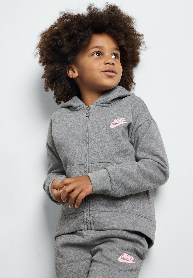 Sudadera gris con cremallera hecha de material de forro polar con un logo de Nike en rosa. Cuenta con una capucha y bolsillos laterales. Combinada con pantalones grises a juego.