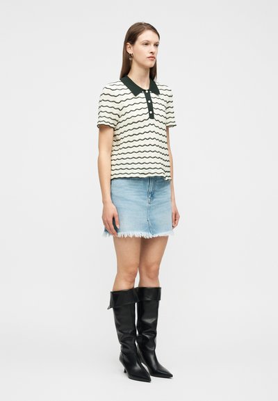 Γυναίκα που φοράει κοντομάνικη ριγέ πόλο μπλούζα με κυματιστά σχέδια, φούστα από ανοιχτόχρωμο ξεφτισμένο τζιν και μαύρες μπότες με ψηλό τακούνι μέχρι το γόνατο, στέκεται μπροστά σε λευκό φόντο.