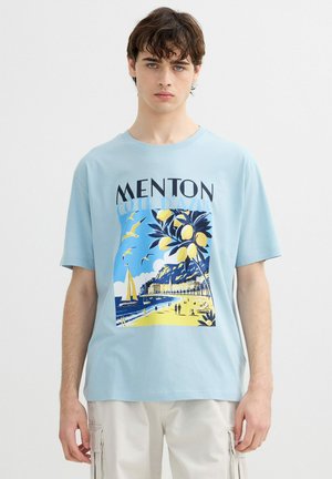 Joven con camiseta azul claro con estampado del litoral de Menton, limones amarillos, gaviotas, velero y gente en la playa.