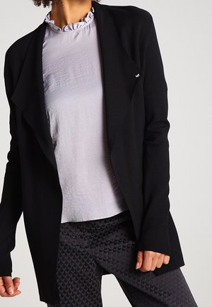 Vrouw met een zwarte blazer met open voorkant, lichtgrijs blouse met gerimpelde kraag en donker patroon broek, staande tegen een effen achtergrond.