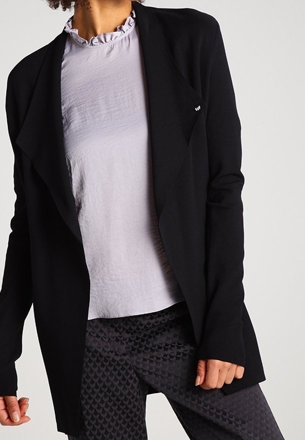 Femme portant un blazer noir ouvert devant, un chemisier gris clair à col volanté et un pantalon foncé à motifs, debout devant un fond uni.