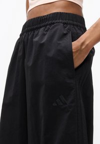 Άτομο που φοράει μαύρο σορτς Adidas με ελαστική μέση, χέρι στην τσέπη, με το λογότυπο να είναι ορατό στον αριστερό μηρό.