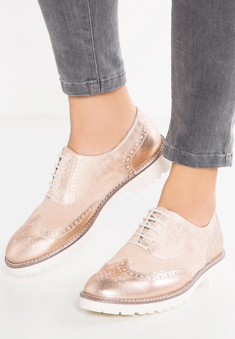 Femme portant des chaussures oxford style brogue en or rose métallique avec des semelles blanches et un jean gris retroussé, debout sur un fond blanc.