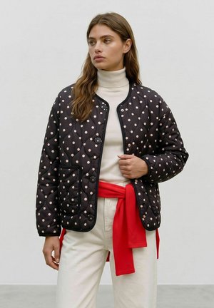 Femme portant une veste matelassée noire à pois sur un col roulé blanc et un pantalon blanc, avec un pull rouge noué autour de la taille.
