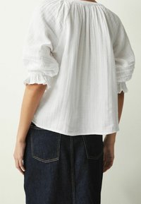Weiße Baumwollbluse mit Puffärmeln, strukturiertem Stoff und Rüschensäumen, kombiniert mit einem dunkelblauen Jeansrock mit Gesäßtaschen.