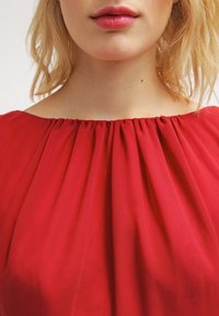 Blusa rossa a pieghe con scollatura ampia. Il tessuto sembra leggero e dalla texture, con morbide pieghe nella zona del collo.