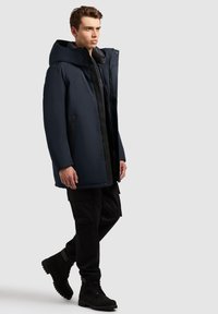 Mand iført en mørkeblå hættetrøje, sorte cargo bukser og sorte snørebåndsstøvler mod en lys grå baggrund.