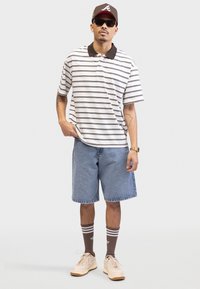 Polo a maniche corte a righe con colletto marrone, pantaloni corti in denim, calze a righe marroni e sneakers beige. Occhiali da sole neri e cappellino marrone.