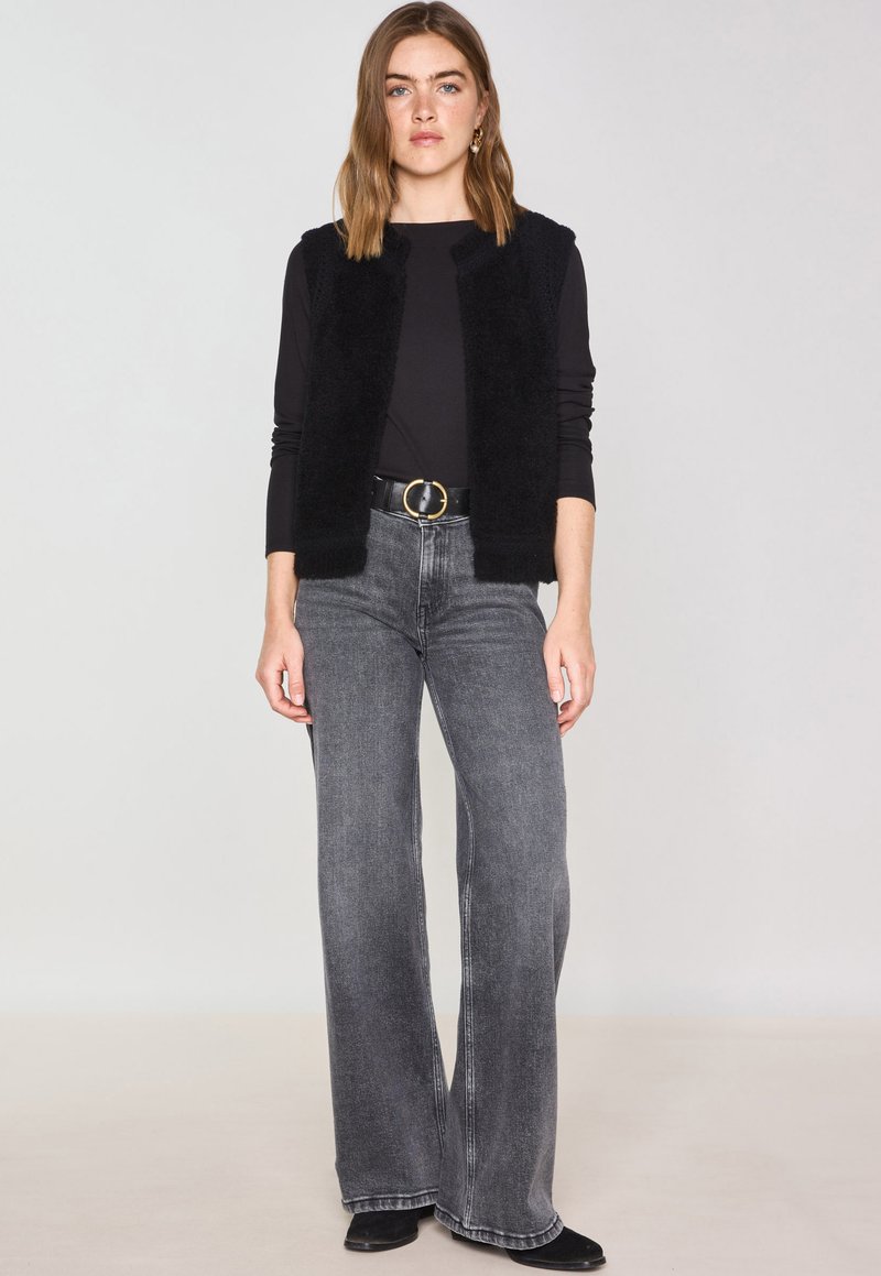 Gilet senza maniche nero realizzato in morbido materiale, abbinato a un top nero e jeans grigi a gamba larga. Presenta un accento con una fibbia rotonda per la cintura.