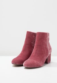 Par de botas de tobillo de terciopelo para mujer en rosa polvo con tacones anchos sobre una superficie blanca.