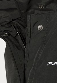 Gros plan sur le tissu noir d'une veste montrant une fermeture éclair partiellement ouverte, des boutons-pression et une partie du logo blanc « DIDRI ».