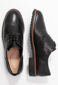 Chaussures noires en cuir à lacets avec une finition lisse, bout arrondi, semelle texturée et un accent marron autour du bord de la semelle.