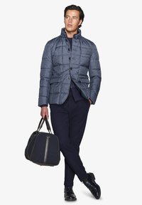 Giacca imbottita blu scuro con colletto e chiusura con bottoni, abbinata a pantaloni scuri e scarpe lucide; tiene una borsa scura con dettagli in pelle.