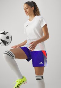 Balta īsām piedurknēm krekls, zili šorti ar sarkaniem akcentiem un baltiem svītrām, baltas ceļgalu garuma zeķes un košas dzeltenas futbola kurpes. Futbola bumba kustībā.